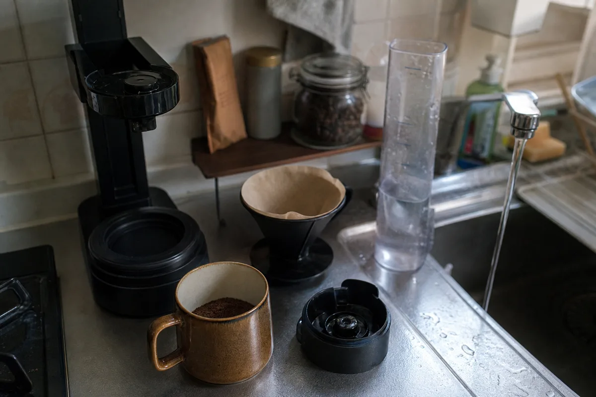 朝のキッチンで豆とマグカップを並べ、ミル付きコーヒーメーカーの置き場所を確認するイメージ